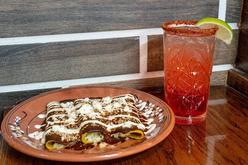 Enchiladas topped with mole, crema and crumbled queso fresco on a hand-painted clay plate, beside a tall red margarita with a chili-salt rim and a lime wedge