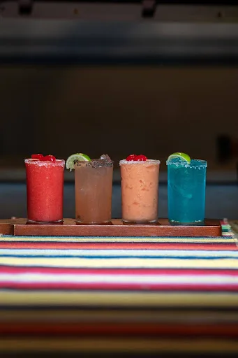 Flight of four frozen margaritas in red, tamarind, peach and blue, with chili-salt rims and lime and cherry garnishes, on a wooden board over a striped serape