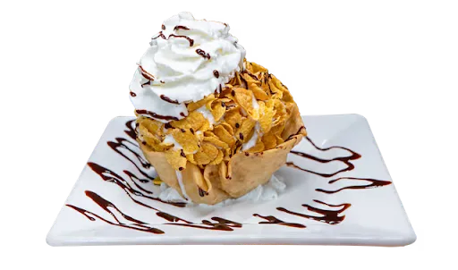 Fried ice cream in a tortilla basket coated in corn flakes, crowned with whipped cream, on a square white plate drizzled with chocolate