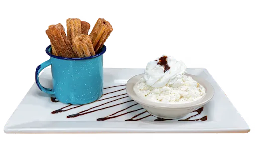 Churros standing in a blue enamel mug beside a bowl of whipped cream, on a white plate drizzled with chocolate
