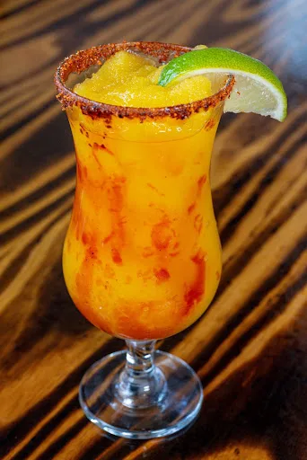 Frozen mango Mangonada in a tall hurricane glass with a chili-salt rim and a lime wedge, on a wooden table