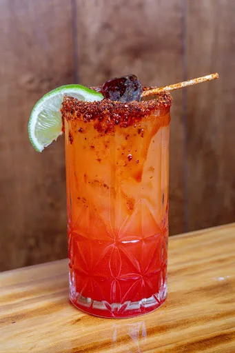 Tall red cocktail with a chili-salt rim, a lime wheel and a tamarind candy straw, on a wooden board