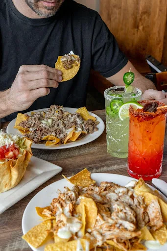 Guest lifting a loaded nacho over shared plates of beef and chicken nachos, with a green and a red cocktail on the table
