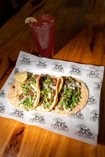 Three street tacos with onion and cilantro on Casa Pirul paper, next to a tall red drink and a lime wedge