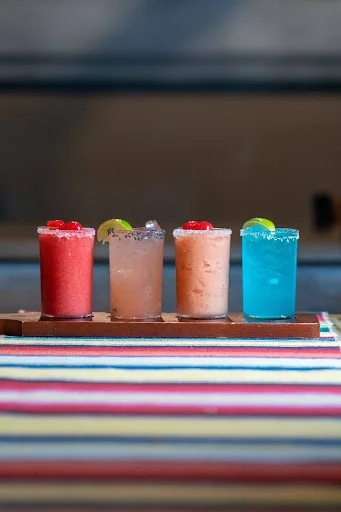 A wooden flight of four frozen margaritas in red, pale yellow, peach and blue, with salted rims and lime wedges